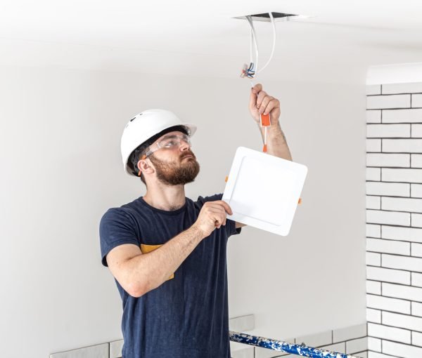 Electrician Builder with beard worker in a white helmet at work, installation of lamps at height. Professional in overalls with a drill on the background of the repair site.