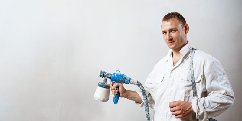 Worker looking at camera, painting wall with spray gun in white color. Copy space.
