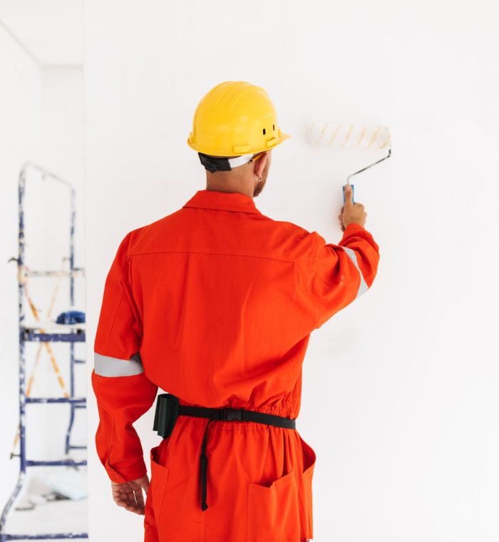 Young foreman standing from back in orange work clothes and yellow hardhat using painting roller in new apartment at work